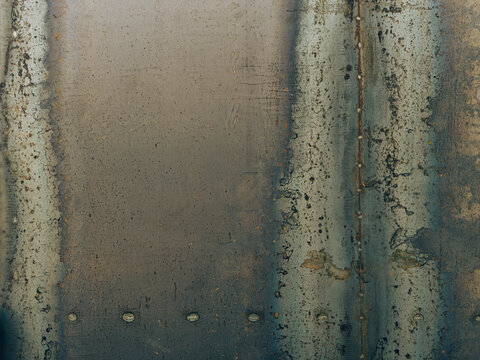 Weathered industrial metal sheet with rustic patches, oxidation, and visible welding seams, creating a dark, textured, and raw metallic background surface