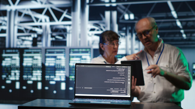 Octogenarian managers in data center, solving tasks on tablet while running scripts on laptop terminal, with adept supervisors amidst servers, accessing databases and manipulating lines of code.
