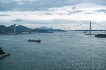 Fototapeta premium Cargo ship sailing under large suspension bridge over calm water with cloudy sky and distant mountains