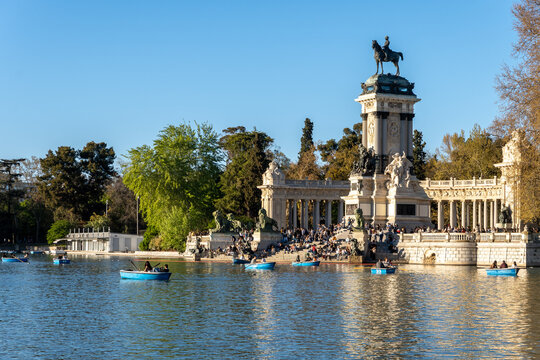 A horizontal scenic shot of the grand Monument to King Alfonso XII and active tourist rowing boats on the lake inside El Retiro Park on a bright sunny day in Madrid, Spain.