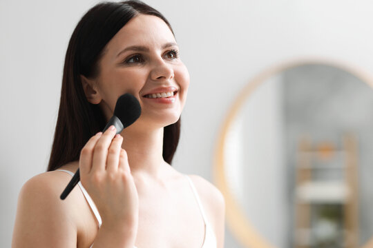 Young woman applying face powder with makeup brush indoors, space for text