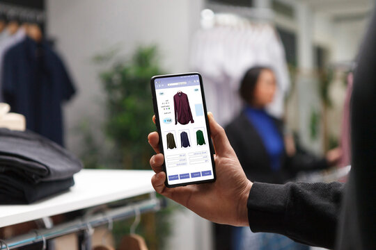 Caucasian customer in a clothes store is holding a smartphone and looking at clothes on a boutique website, male shopaholic is perusing clothing items in a mall using a cellphone.