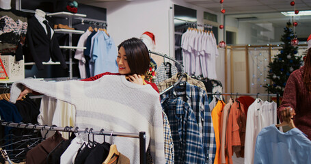 Fototapeta premium Smiling client in Christmas ornate fashion shop looking to purchase comfortable clothes on clearance, taking advantage of reduced prices during holiday season promotional sales.