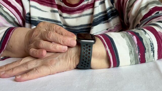 Close-up of an elderly woman's hands wearing a smartwatch. The device displays heart rate information. The woman has wrinkled skin and is wearing a patterned shirt