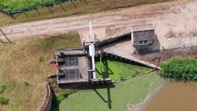 Aerial view of Islington Pumping Station featuring the sluice gate, pipes, and a small brick building by the algae-covered water in King's Lynn, England, United Kingdom.