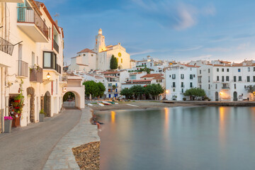 Obraz premium Early morning view of the waterfront and traditional white buildings in Cadaques, Spain. Calm bay waters reflect the first light of dawn along the Mediterranean coast.