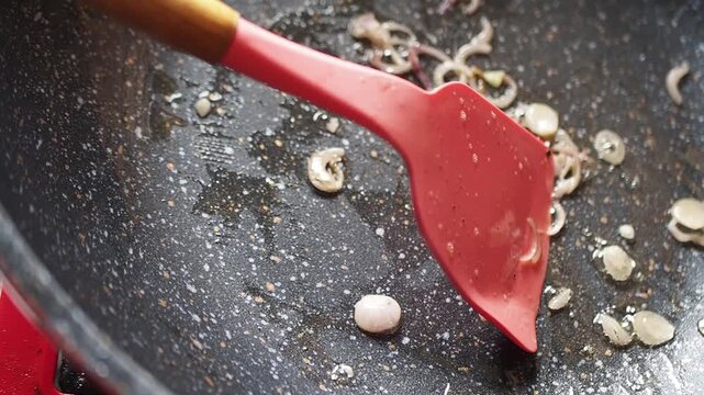 Close-up of a red silicone spatula stirring sliced shallots in a hot, oily non-stick frying pan, preparing food for cooking.