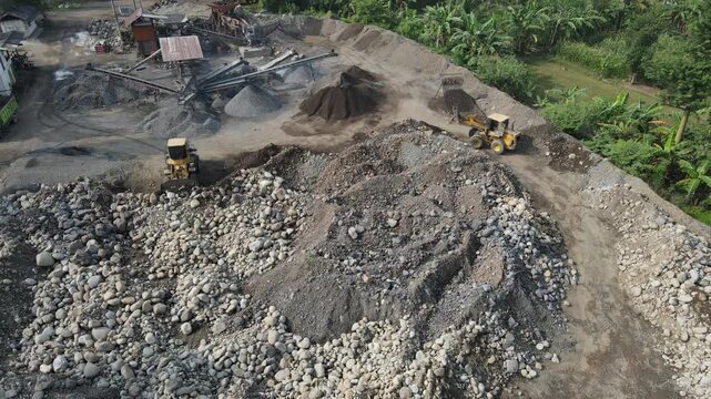 Aerial view of heavy machinery operating at a sand and stone quarry with piles of gravel and lush greenery in Patikraja, Central Java, Indonesia.