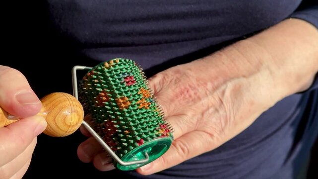 Close-up hand massage using needle roller performed by woman caregiver on palm and fingers, green spiked roller and wooden handle