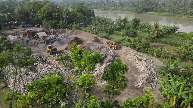 Aerial view of a stone quarry with heavy machinery, piles of rocks and lush green trees near a river in Patikraja, Central Java, Indonesia.