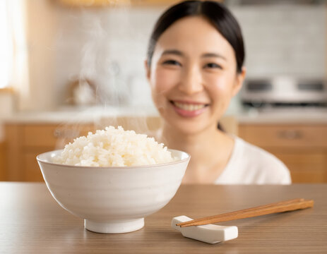 Smiling woman looking at a steaming bowl of white rice, chopsticks resting on a white ceramic holder, wooden table, home kitchen, healthy Japanese cuisine.