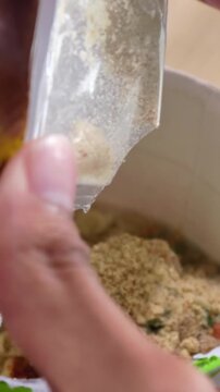 Close up of a hand pouring dry powder seasoning from a sachet into an instant cup noodle bowl.