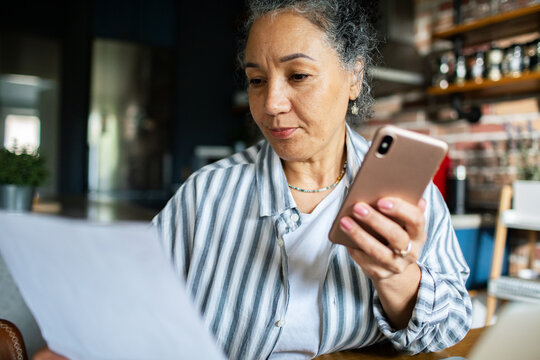 Mature woman reviewing bills with smartphone at home kitchen