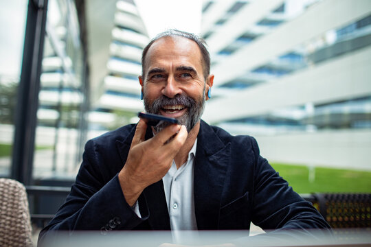Smiling mature businessman using smartphone voice assistant at outdoor office campus