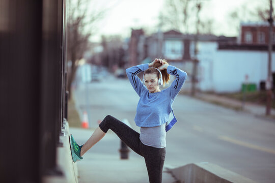 Young woman runner stretching with headphones on city sidewalk