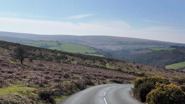 Dartmoor Devon England UK.  27.03.2026. Video. A paved road curves through a picturesque landscape of green fields,  on Dartmoor Devon with dense forests, and rolling hills under a bright blue sky