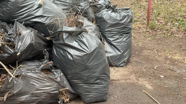 Black garbage bags piled by fence, on roadside near small green trash can, scattered leaves and gravel, neglected urban corner