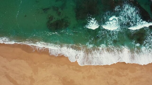 Aerial drone shot of turquoise ocean waves rolling onto sandy beach shoreline. Scenic coastal landscape with sea foam and clear water texture from above