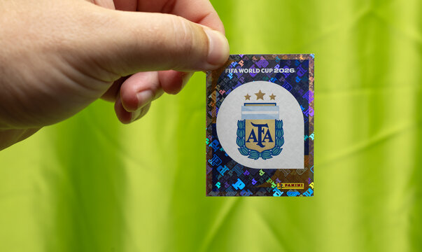 person holding a shiny holographic panini sticker of the argentina national football team crest for the fifa world cup 2026 on a bright green background