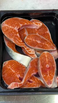 Middle frame of retail seafood sequence showing multiple fresh salmon steaks displayed in black plastic tray at counter center, overhead bright lighting creates glossy wet sheen on orange