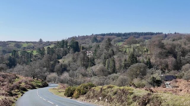 Dartmoor Devon England UK.  27.03.2026. Video. A paved road curves through a picturesque landscape of green fields,  on Dartmoor Devon with dense forests, and rolling hills under a bright blue sky