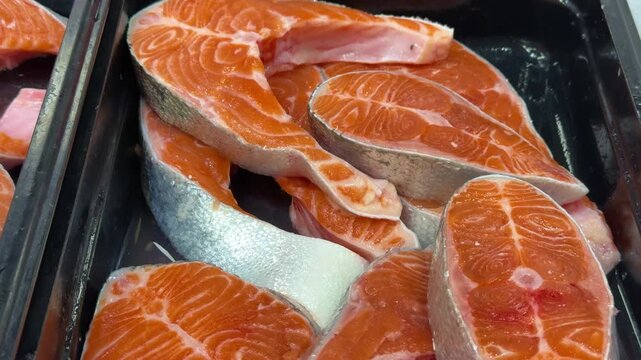 Middle frame of retail seafood sequence showing multiple fresh salmon steaks displayed in black plastic tray at counter center, overhead bright lighting creates glossy wet sheen on orange