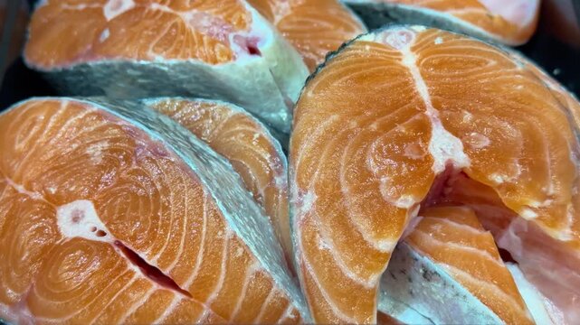 Middle frame of retail seafood sequence showing multiple fresh salmon steaks displayed in black plastic tray at counter center, overhead bright lighting creates glossy wet sheen on orange
