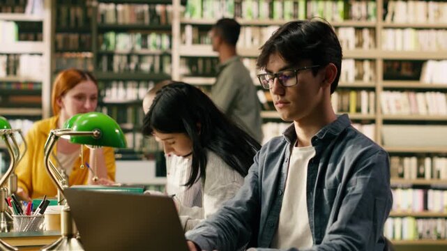 Asian student with glasses working on laptop at university library table, reading and typing information on computer during self study session, Focused academic environment, with academic research.