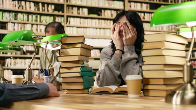Fatigued young woman covering mouth yawning over open book at library desk, surrounded by books stacks and study materials during academic preparation.