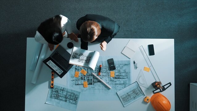 Time lapse of skilled project manager holding project plan and explain house design to architect engineer at meeting table with equipment, ruler, safety helmet and laptop scatter around. Alimentation.