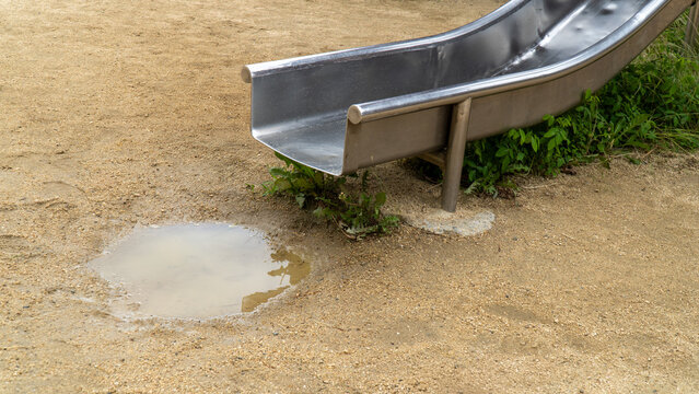 Metal Playground Slide Bottom with a Puddle on Sandy Park Ground