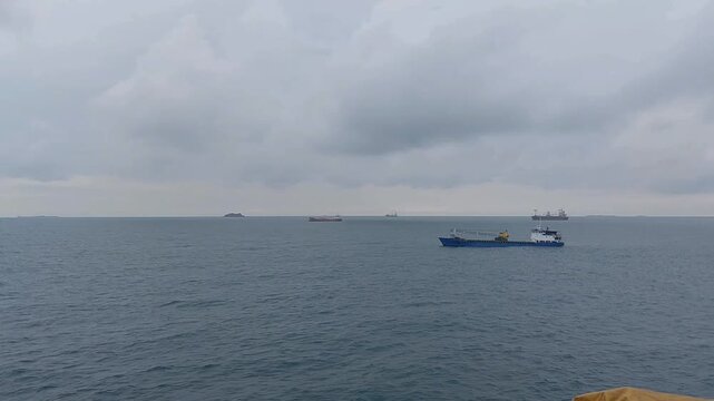 Wide shot of the ocean near Makassar, Indonesia, viewed from a moving ship. Static camera captures a blue cargo vessel crossing calm waters under a cloudy afternoon sky.