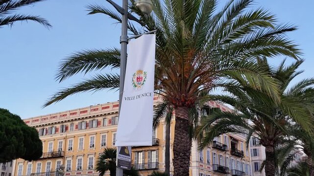 Static medium shot of the white Ville de Nice flag with the city's official coat of arms hanging on a lamppost next to palm trees at sunset.