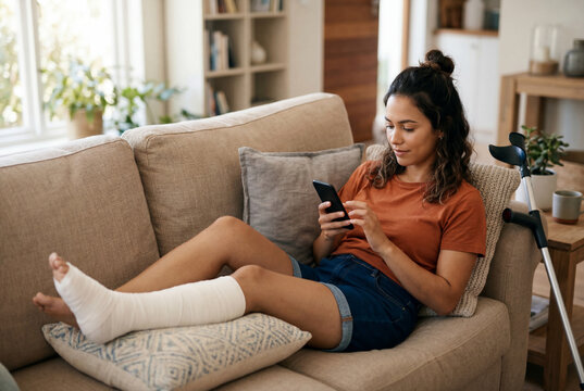 Woman with broken leg in cast relaxing on couch using smartphone