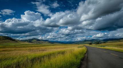 Fototapeta premium Scenic Landscape of Open Road Through Lush Green Fields Under Dramatic Blue Sky with Fluffy White Clouds in the Background of Majestic Mountains