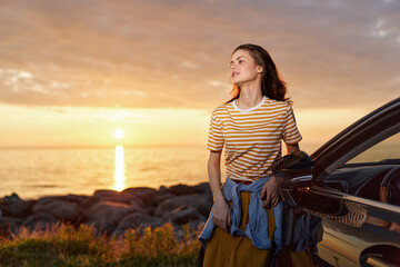 Fototapeta premium A young woman stands beside a car at sunset, wearing a striped shirt and denim jacket tied around her waist, looking toward the glowing horizon and calm coastline.