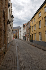 Naklejka premium Cobblestone street view of Heilige-Geeststraat in Bruges, Belgium, lined with historic buildings under a cloudy sky.