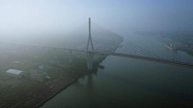 Aerial drone approaches the silhouette of the Tampico suspension bridge through dense fog at dawn, with low moody light revealing the Panuco River glowing faintly through the mist