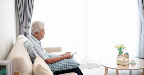 Fototapeta premium Senior man sitting on a sofa and using a remote control in a bright living room. Retirement lifestyle, home leisure, technology use, independent living, and active aging concept with copy space.