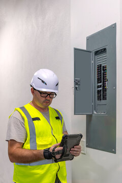 Professional electrician using digital tablet to inspect or log data next to open wall mounted electrical breaker panel