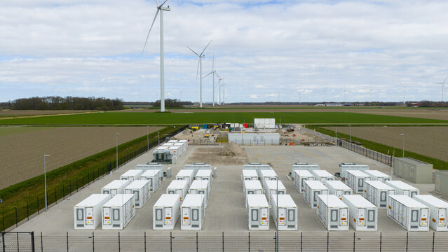 Dronten, Netherlands - 04 April 2026: Aerial view of a battery energy storage system facility with rows of white containers and wind turbines in the background.