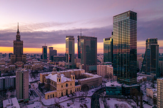 Warszawa, Poland - 05 January 2026: Aerial view of Plac Grzybowski and All Saints Church surrounded by modern skyscrapers and the Palace of Culture and Science under a purple sunset sky.