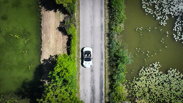 Succieu, France - 13 June 2026: Aerial view of a white convertible car driving on a narrow road between two ponds with lily pads and green vegetation.