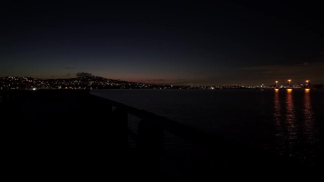 Gimbal close-up panning shot of the Cabrillo Beach jetty with the Port of Los Angeles and Palos Verdes in the distance during night in San Pedro, California. 4K