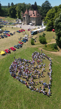 Succieu, France - 13 June 2026: Aerial view of Chateau de Milliassieres with a large crowd of people forming a heart shape on the grass during a car event under a clear sky.