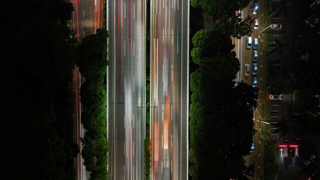 Cilandak, Indonesia - 10 April 2026: Aerial view of long exposure light trails on a highway at night, surrounded by lush trees and a parking lot in Lebak Bulus.