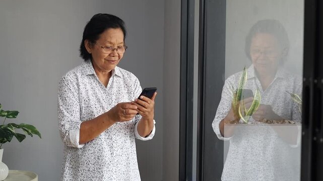 Happy elderly Asian woman standing by window, holding cellphone in hands, elderly lady contacts her family