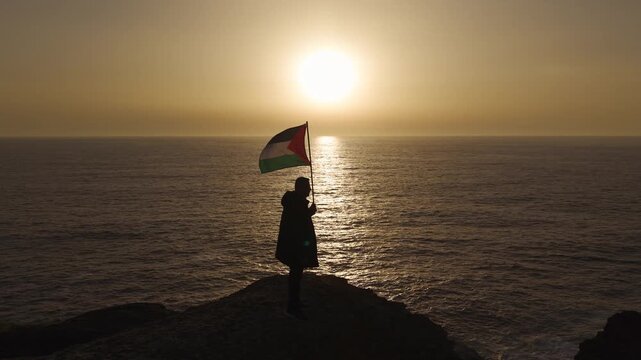 Silhouetted Man Holding A Palestinian Flag Against A Sunset Ocean Background In Spain. - aerial shot