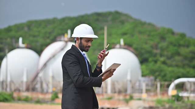 Engineers wearing safety gear, including hard hats examining survey a large blueprint tablet standing industrial facility gas or oil refinery engaged in a job requires high safety standards concept.	