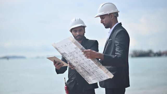 Engineers wearing safety gear, including hard hats examining survey a large blueprint tablet standing industrial facility gas or oil refinery engaged in a job requires high safety standards concept.	